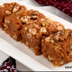 A close-up image of a traditional sweet dish called "sohan halwa," showcasing its rich golden-brown color and garnished with chopped pistachios and almonds, served on a decorative plate.